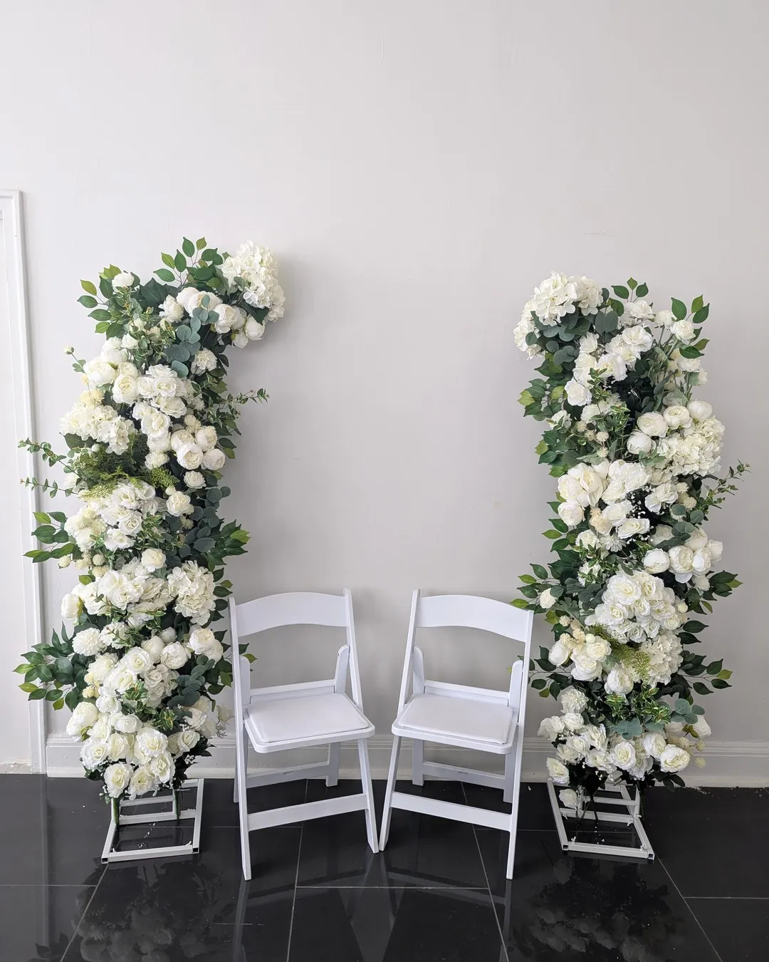 Two columns of white flowers and greenery framing a pair of white folding chairs