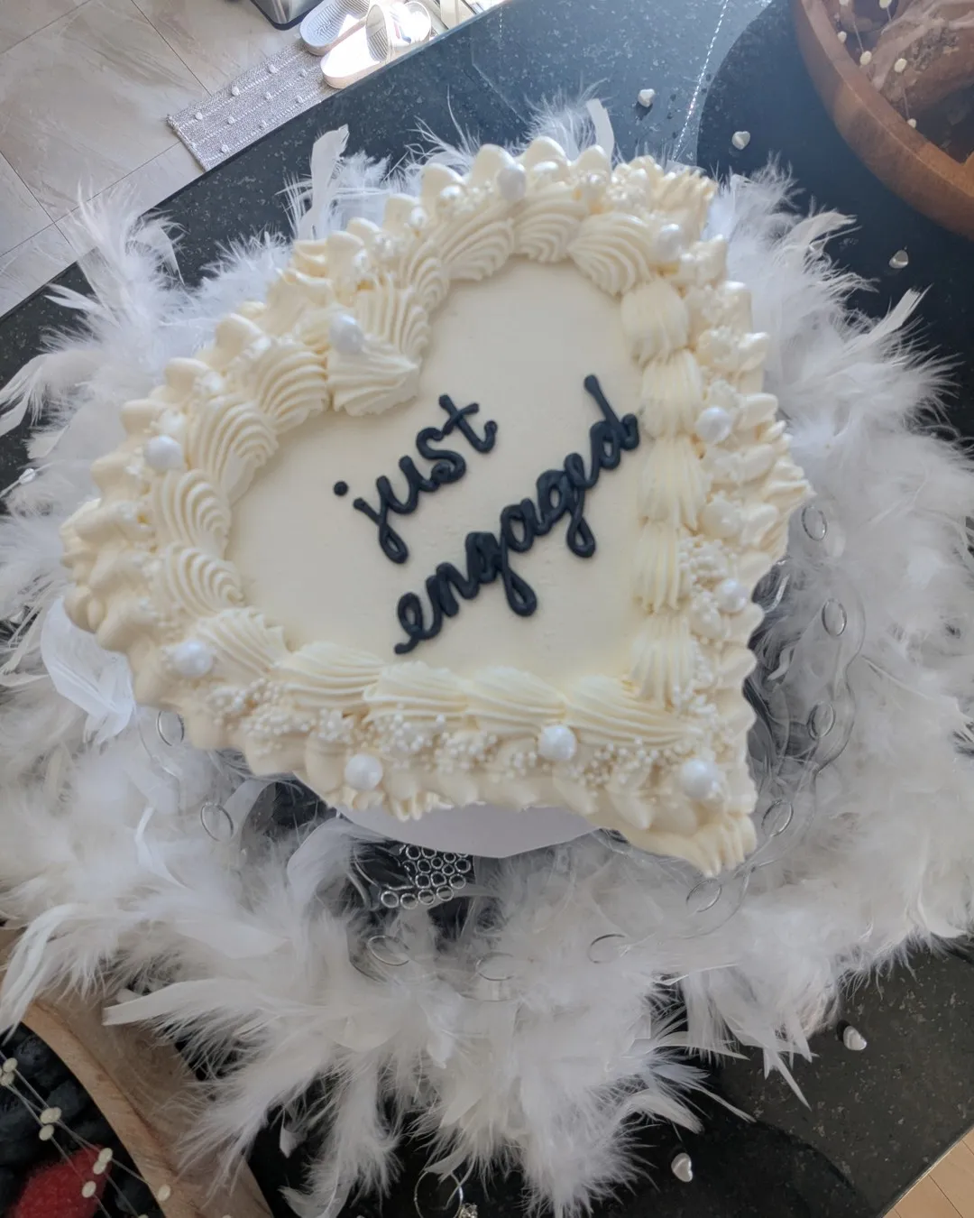 Heart-shaped cream cake reading "just engaged" on a ring of white feathers
