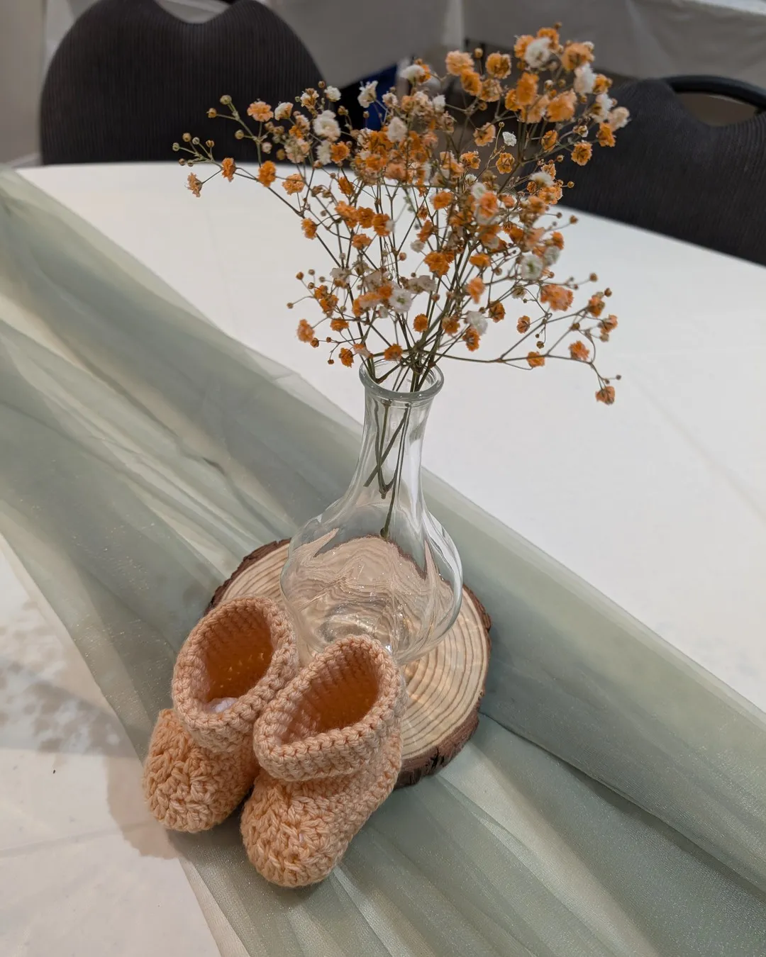Knitted peach baby booties beside a bud vase of dried flowers on a sage runner