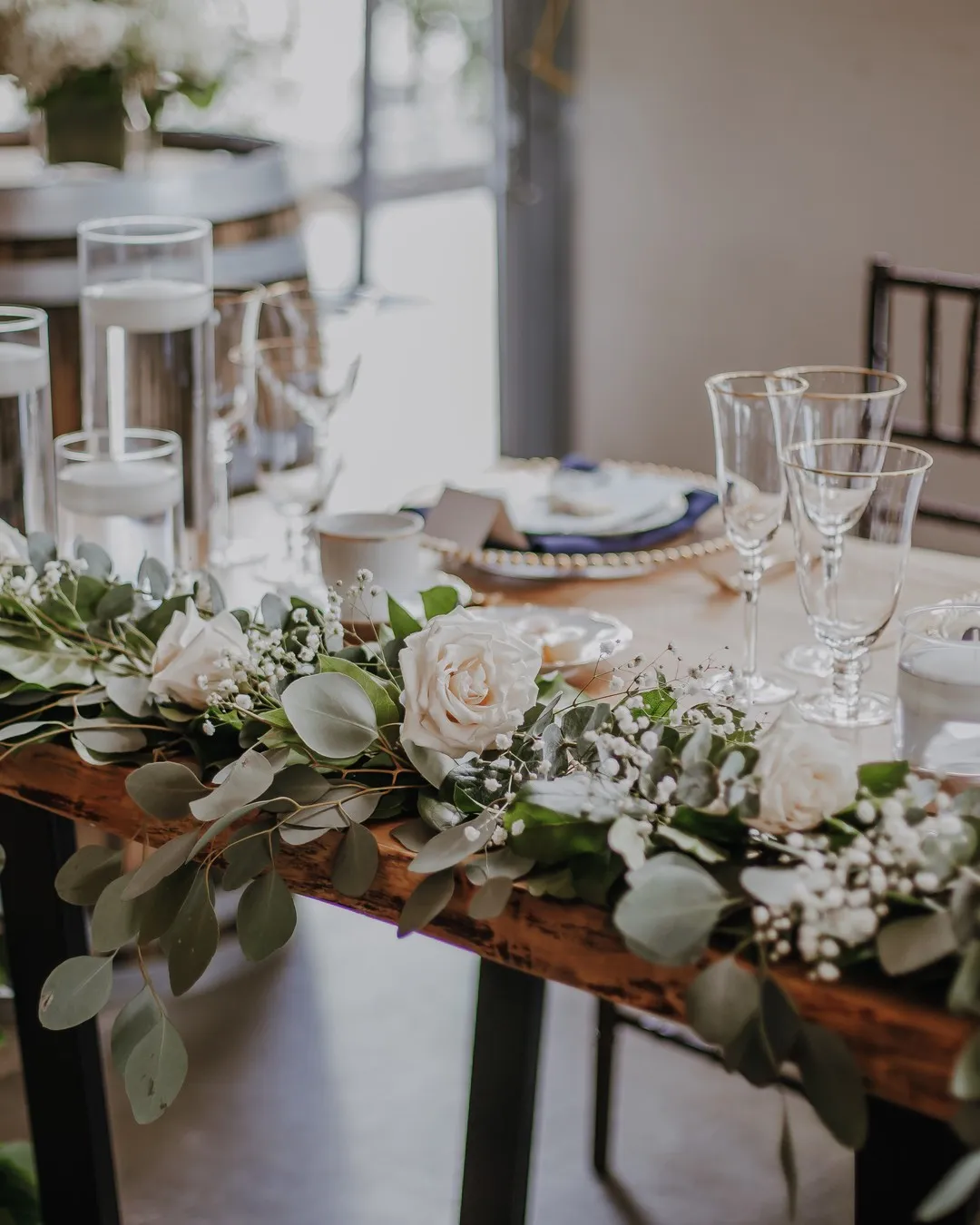 Long wooden table with a low garland of white roses and eucalyptus, floating candles and crystal glassware