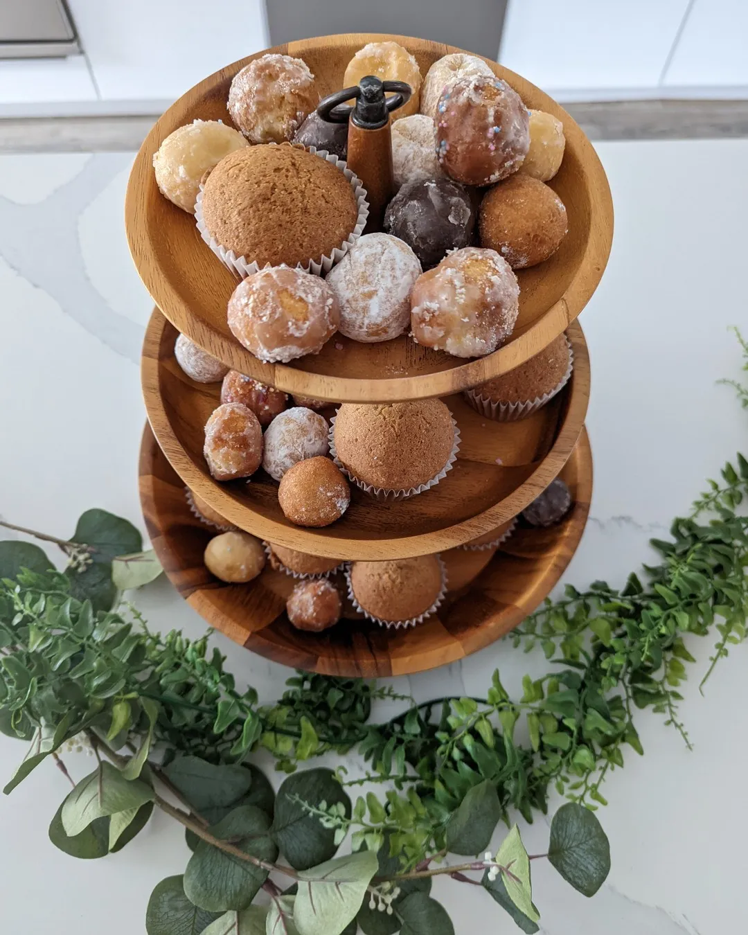 Three-tier wooden stand of muffins and sugared doughnut holes on a eucalyptus garland