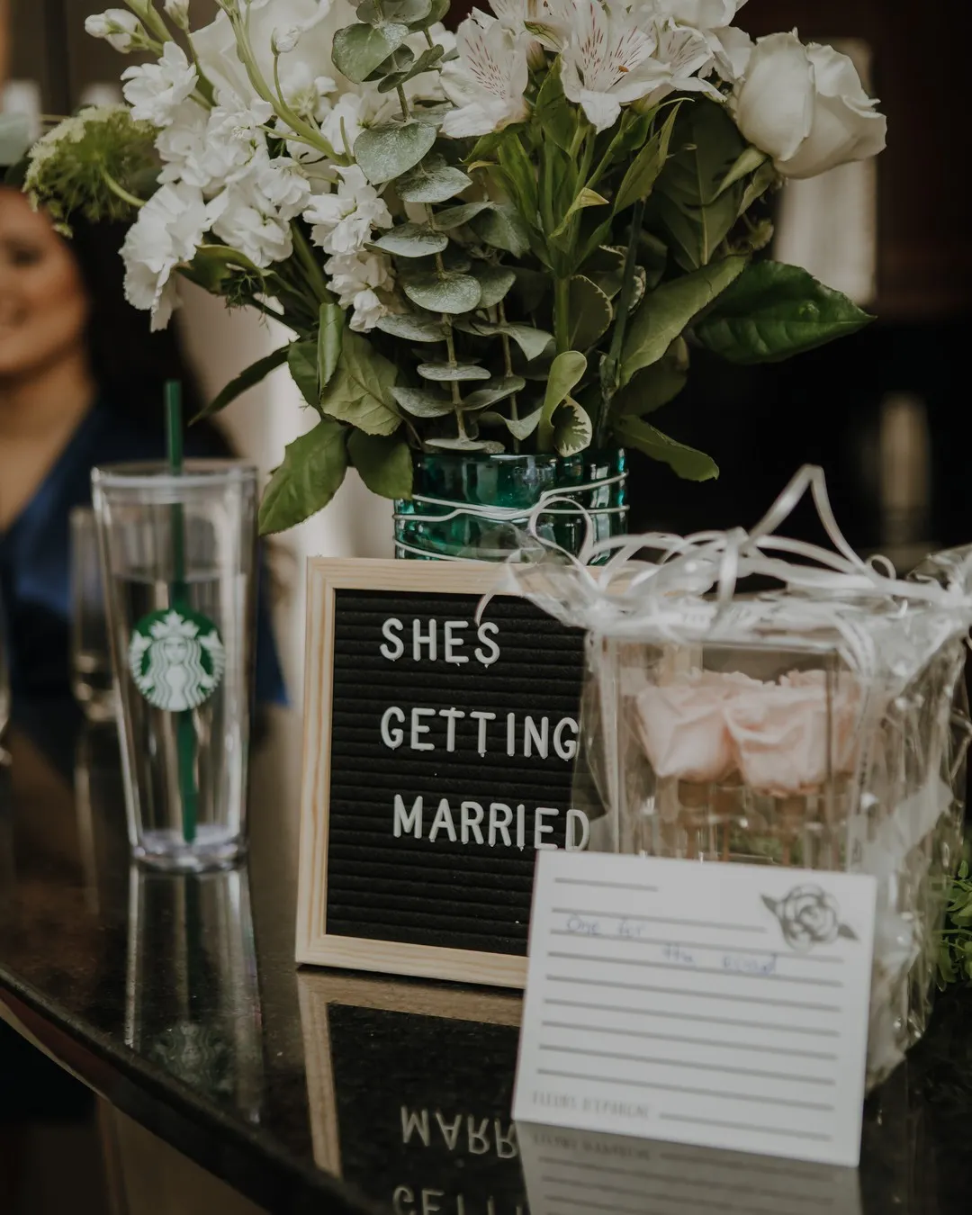 Letter board reading "She's getting married" beside white flowers, a ribbon-tied box of blush roses and a lined guest card