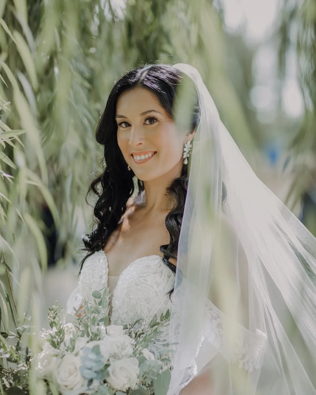 Nicole in her wedding gown and veil among willow branches, holding a white rose bouquet